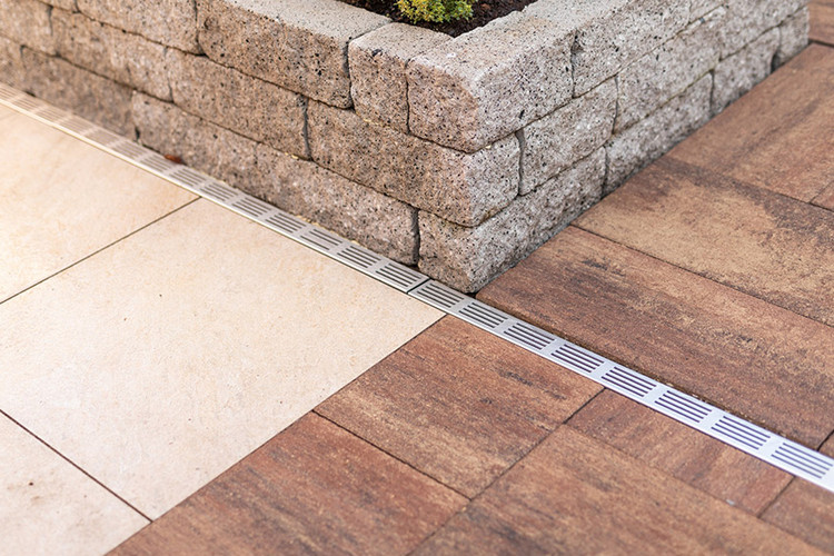 Detailansicht einer Terrasse mit hellen und dunklen Steinplatten, Entwässerungsrinne und angrenzender Palisade aus Naturstein.