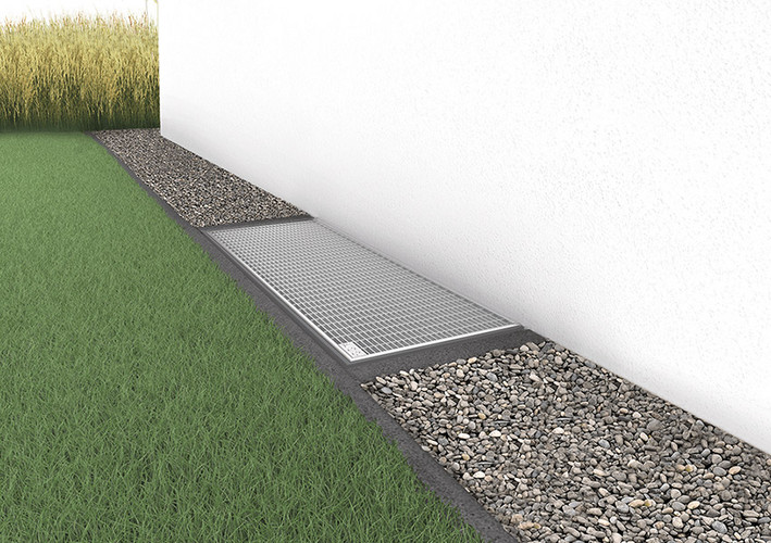 Außenbereich mit Gras, einem Lichtschacht in einem schmalen Kiesbett entlang einer Hauswand.