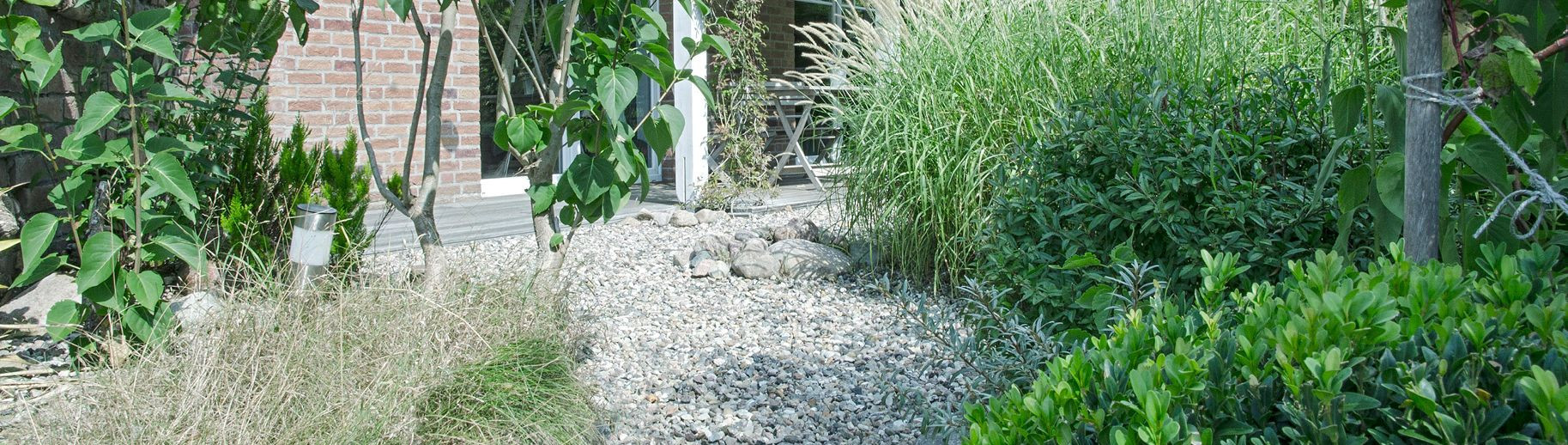 Garten mit Kiesweg, umgeben von grünen Pflanzen und Sträuchern, im Hintergrund ein Backsteingebäude und eine Sitzgruppe.