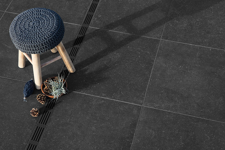 Dunkle Terrassenplatten mit schmaler Entwässerungsrinne, darauf ein Holzhocker mit blauem Sitzpolster.