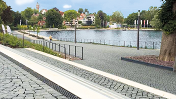 Gepflasterter Uferbereich mit Entwässerungsrinne, Blick auf See und umliegende Häuser im Hintergrund.