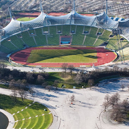 Außenansicht des Olympiastadions in München, mit seiner ikonischen, transparenten Zeltdachkonstruktion. Das Stadion hat eine ovale Form und ist von geschwungenen, lichtdurchlässigen Dachflächen umgeben. Die Architektur ist innovativ und futuristisch, und im Hintergrund sind grüne Parklandschaften und städtische Gebäude sichtbar.