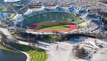 Außenansicht des Olympiastadions in München, mit seiner ikonischen, transparenten Zeltdachkonstruktion. Das Stadion hat eine ovale Form und ist von geschwungenen, lichtdurchlässigen Dachflächen umgeben. Die Architektur ist innovativ und futuristisch, und im Hintergrund sind grüne Parklandschaften und städtische Gebäude sichtbar.