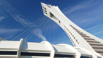 Außenansicht des Olympiastadions in Montreal, mit seiner markanten, schrägen Turmstruktur, die über das Stadion hinausragt. Das Stadion hat eine ovale Form und ist von einem weitläufigen Dach bedeckt. Die Architektur ist modern und innovativ.