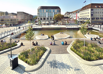 Urbaner Platz mit Wasserfläche, begehbaren Inseln, Holzdecks und Menschen, die entspannen.