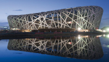 Außenansicht des Olympiastadions in Peking mit seiner einzigartigen, verschachtelten Stahlkonstruktion. Das Stadion hat eine ovale Form und ist von einem komplexen Netz aus ineinander greifenden Streben umgeben. Die Fassade ist modern und futuristisch. Das Stadion liegt direkt am Wasser.