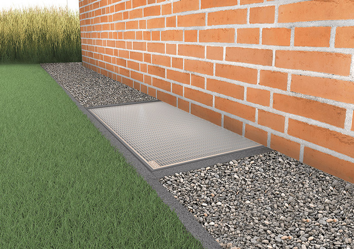 Lichtschacht an einer Klinkerfassade, schmales Kiesbett entlang der Hauswand umgeben von Gras.