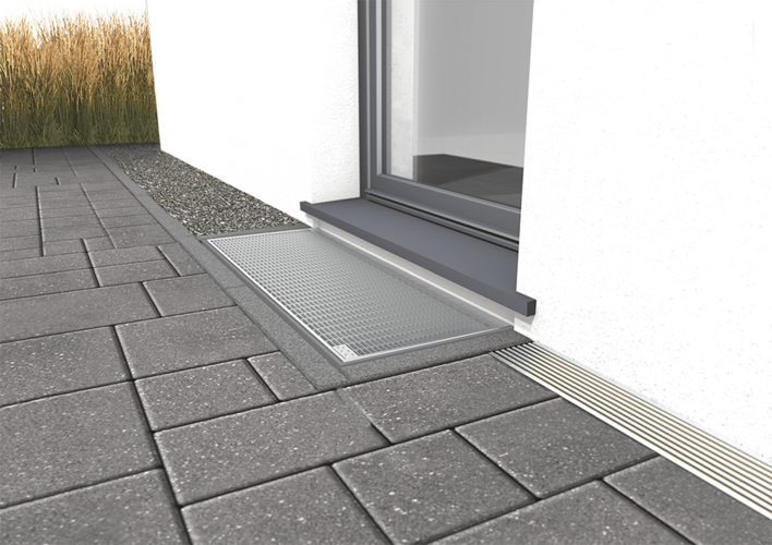 Lichtschacht vor einer Terrassentür mit Schwelle, Entwässerungsrinne, umgeben von Pflastersteinen und einem Kiesstreifen.