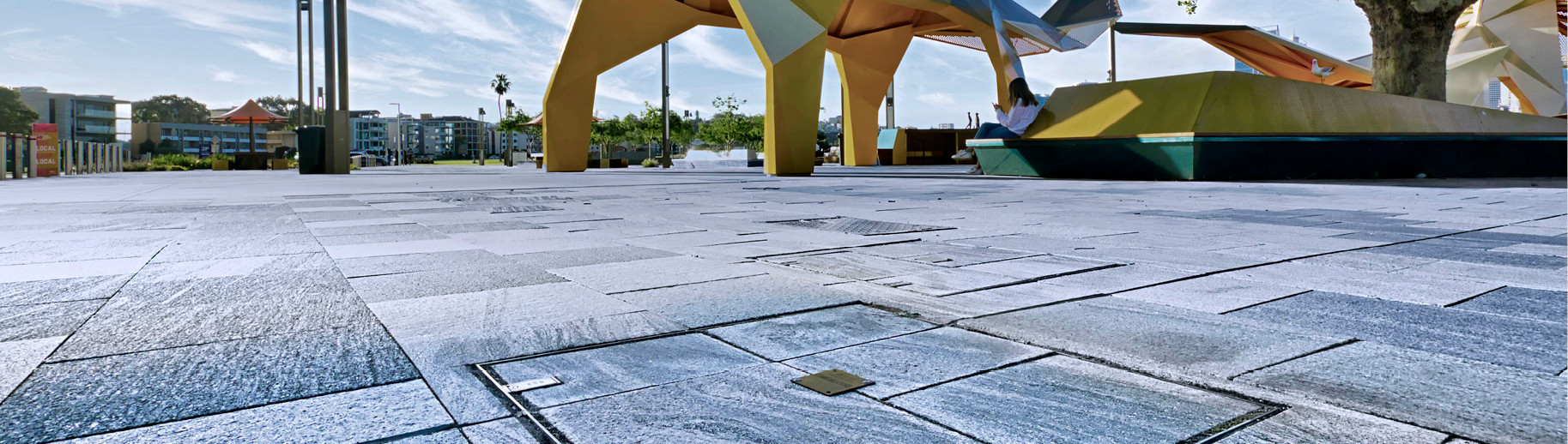 Moderner Platz mit Natursteinplatten, großen geometrischen Skulpturen, Gebäuden und Bäumen im Hintergrund.