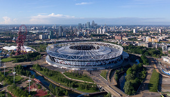 Das Olympiastadion in London, ein großes, ovales Sportstadion mit markanten weißen Stahlträgern und einem offenen Dach. Die Tribünen bieten Platz für zahlreiche Zuschauer. Im Hintergrund sind die Skyline von London und grüne Parkflächen sichtbar.