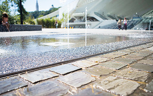 Innerstädtischer Wasserplatz mit Schlitzrinne, vor Gebäuden und mit Personen im Hintergrund