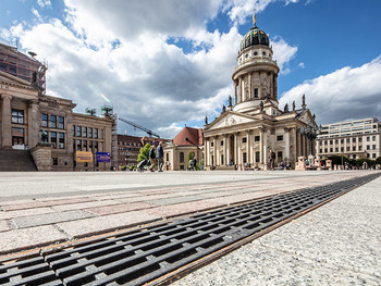 Eine Entwässerungsrinne führt den Blick entlang eines großen Platzes, auf dem ein Dom steht, im Hintergrund sind Menschen.
