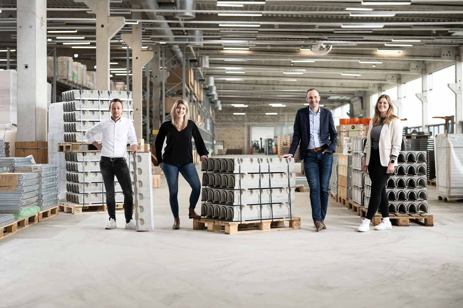 Vier Personen stehen in einer großen Lagerhalle zwischen Paletten mit Entwässerungsrinnen und Bauelementen, lächeln und sehen den Betrachter an.