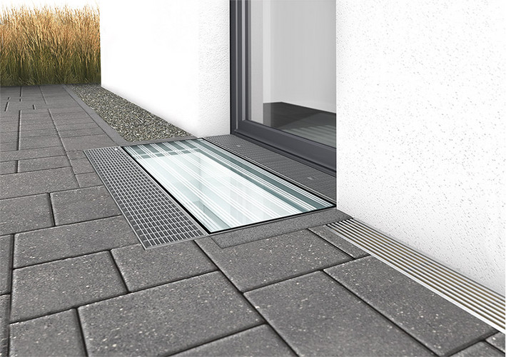 Lichtschacht mit Glasabdeckung vor einer Terrassentür, rechts eine Entwässerungsrinne, links ein schmaler Kiesstreifen, umgeben von Pflastersteinen.