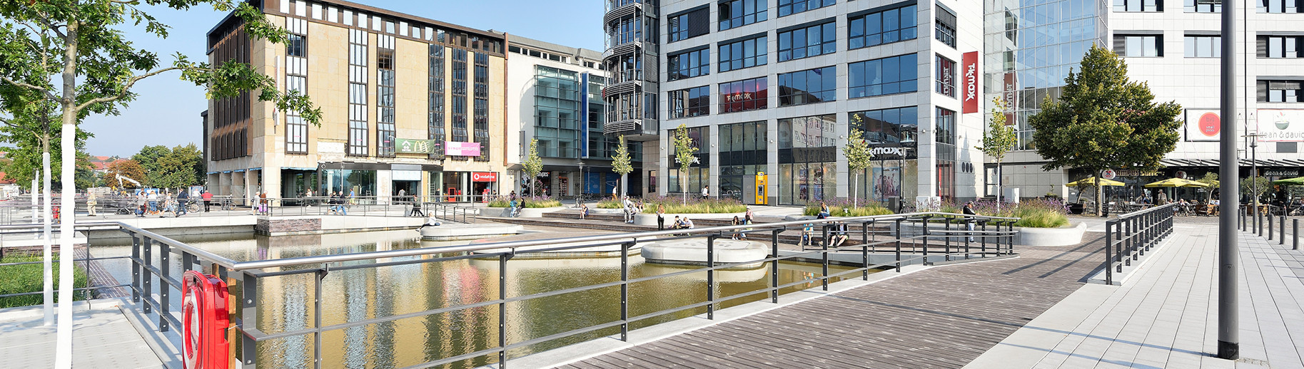 Urbaner Platz mit Wasserfläche, Holzsteg und begehbaren Inseln, umgeben von Bürogebäuden, Geschäften, Bäumen und Grünflächen.