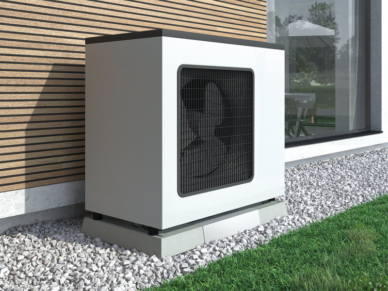 Eine Wärmepumpe an einer holzverkleideten Hauswand, umgeben von einem schmalen Kiesbett, das von Gras gesäumt wird.
