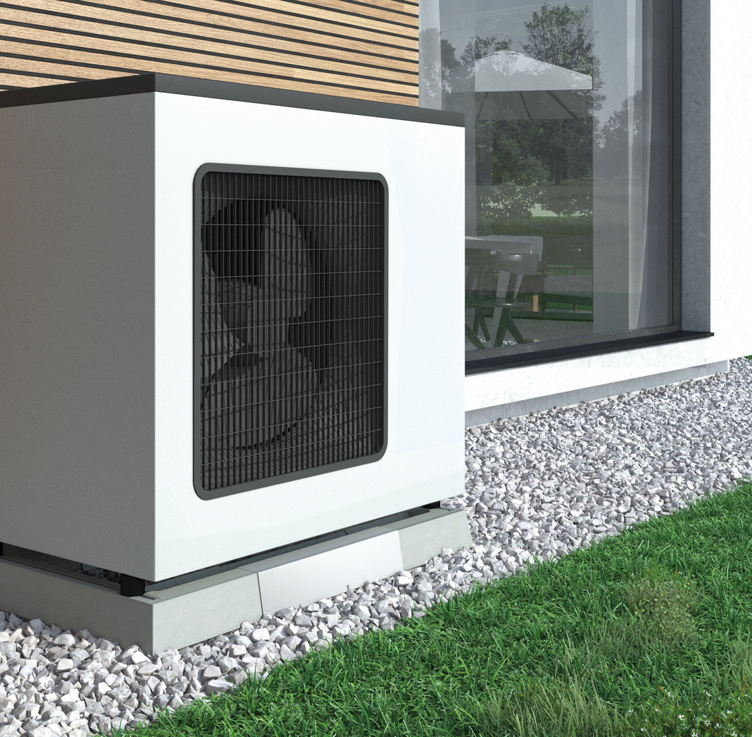 Eine Wärmepumpe an einer holzverkleideten Hauswand, umgeben von einem schmalen Kiesbett, das von Gras gesäumt wird.
