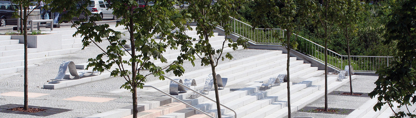 Gestufte Treppenanlage mit integrierten Sitzbereichen, Handläufen und Bäumen im öffentlichen Freiraum.