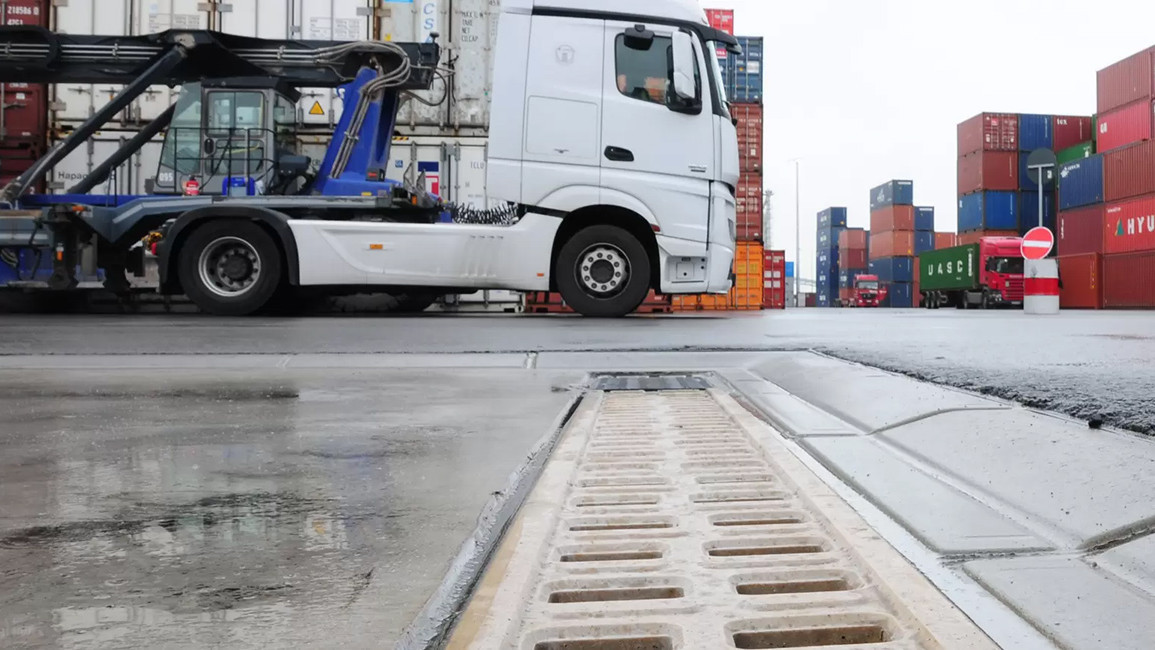 Industrie und Logistik LKWs und Container stehen auf einem Hafenplatz mit Entwässerungsrinne im Vordergrund.