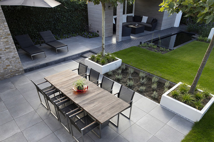 Moderne Gartenterrasse mit großem Esstisch, schwarzen Stühlen, zwei Liegen, Grünflächen und kleinem Wasserbecken.